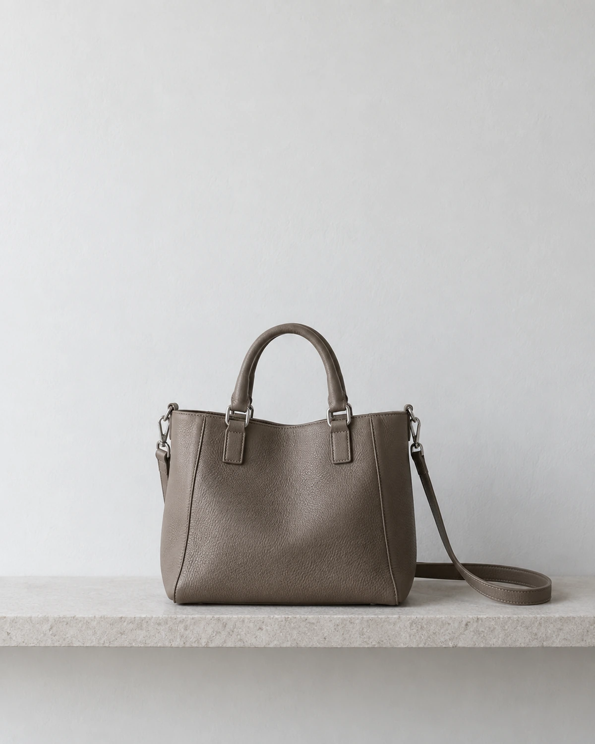 An unbranded dark taupe handbag with a shoulder strap resting on a pale stone shelf.